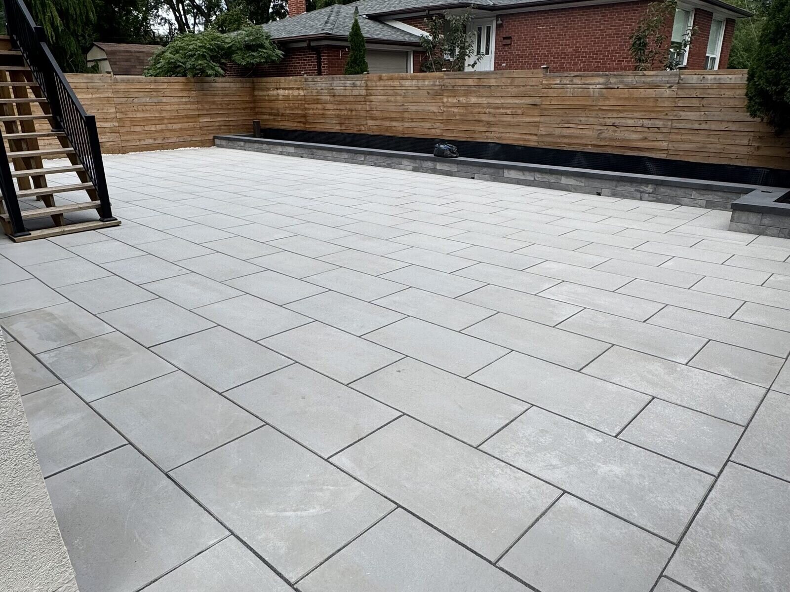 Techo-Bloc Blu 60 large-format slab patio in a North York backyard with a charcoal retaining curb along a horizontal cedar fence and black steel stairs